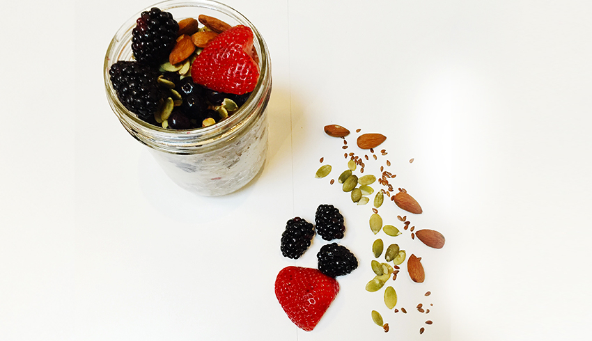 A top view of a mason jar oatmeal and fruit parfait and some of its ingredients, including a strawberry, blackberries, almonds, pumpkin seeds, and flaxseeds, scattered in front of the jar.